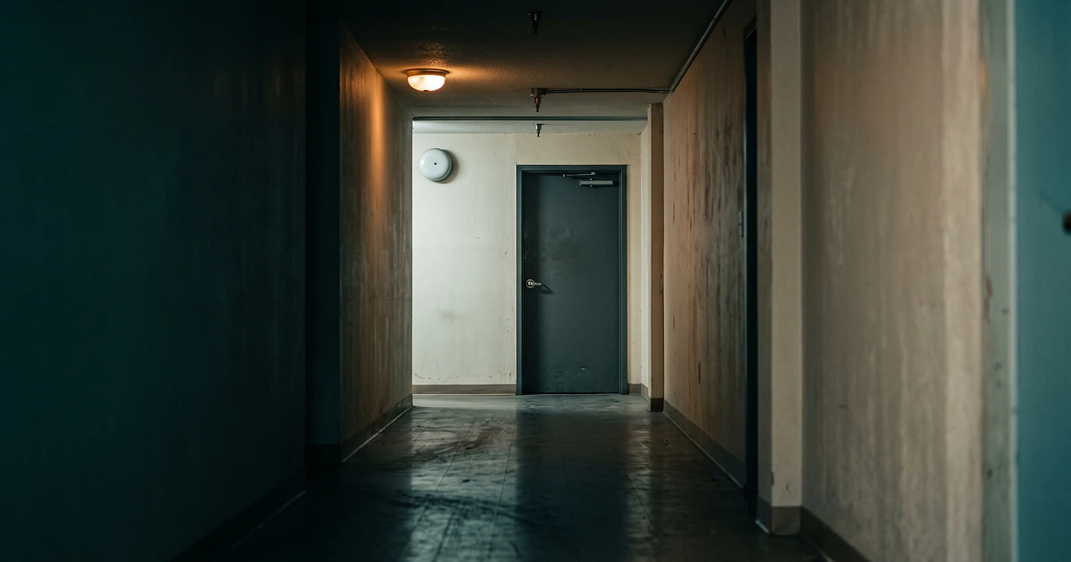 Verlassener Innengang eines kleinen Gewerbegebäudes bei Nacht, niemand zu sehen, ein schlichtes Alarmgehäuse hoch an der kahlen Wand, eine Deckenleuchte, am Ende eine geschlossene Stahltür.
